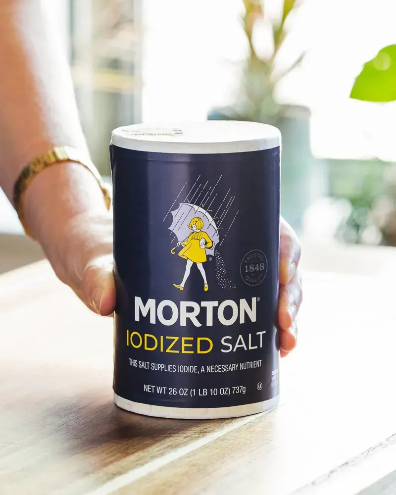 Hand holding a canister of Morton Iodized Salt on a wooden table.